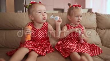 两个小双胞胎姐妹穿着迷人的红色连衣裙，光着脚坐在房间里，兴高采烈地吹着肥皂泡。